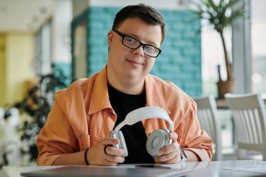 Down sendromlu bir adam bir kafede oturuyor, kulaklık takıyor, gülümsüyor ve kameraya bakıyor..