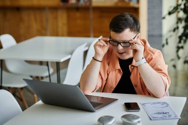 A man works remotely on a laptop at a cafe, adjusting his glasses while focused on his screen. clipart