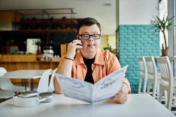 Человек с синдромом Дауна сидит в кафе и смотрит в меню, разговаривая по телефону..