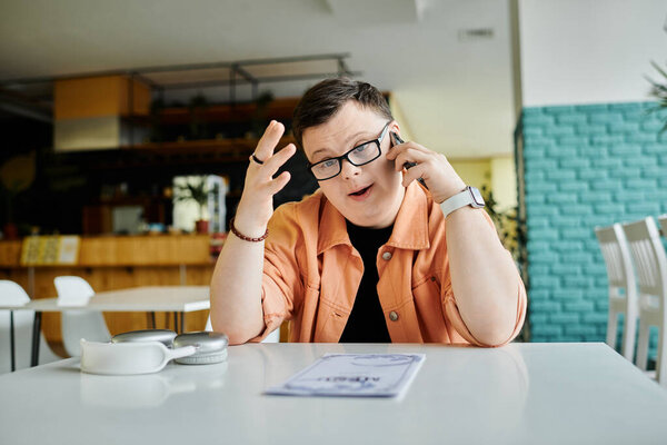A man with Down Syndrome, wearing glasses and a peach jacket, sits at a cafe table, speaking on his phone and looking at the menu.