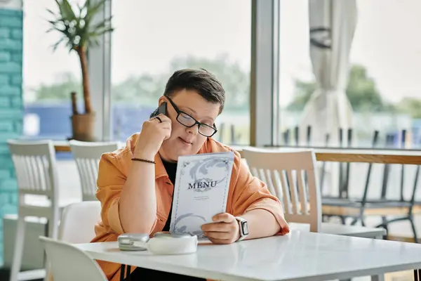 stock image A man with Down Syndrome sits in a cafe, reviewing the menu while holding a phone to his ear.