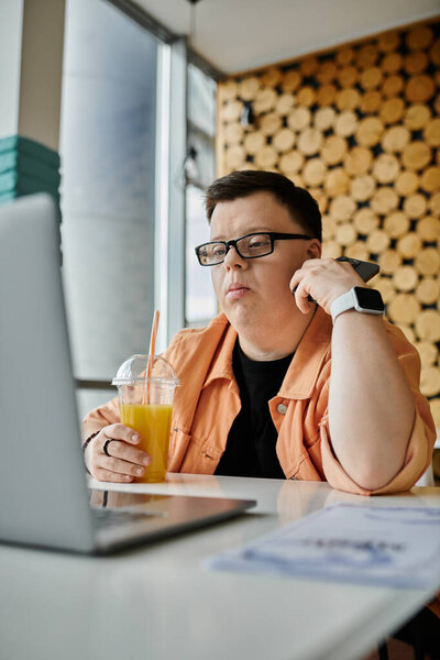A man with Down syndrome sits in a cafe, focused on his laptop, a glass of orange juice in his hand.