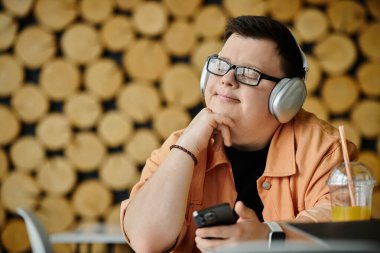 A man with Down syndrome, wearing headphones, sits in a cafe, calmly holding his phone while working remotely. clipart