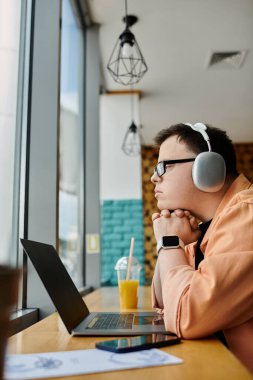 Down sendromlu, kulaklık takan bir adam bir kafe masasında oturuyor ve yanında bir içkiyle laptopuyla çalışıyor..