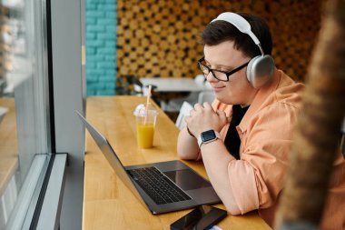 Down sendromlu bir adam bir kafede oturuyor, dizüstü bilgisayarıyla çalışıyor, kulaklık takıyor ve önünde içki içiyor..