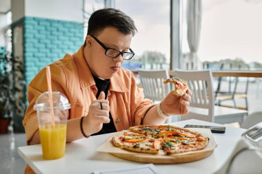 Down sendromlu bir adam bir kafede oturmuş bir bardak meyve suyuyla lezzetli bir dilim pizzanın tadını çıkarıyor..
