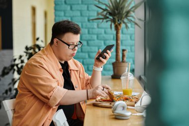 Down sendromlu bir adam kafeterya masasında oturmuş elinde akıllı bir telefonla pizzanın tadını çıkarıyor..