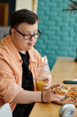 Down sendromlu bir adam bir kafe masasında oturmuş bir dilim pizzanın ve içeceğin tadını çıkarıyor..