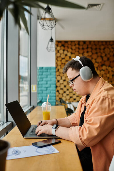 A man with Down syndrome works on a laptop in a cafe, headphones on, with a glass of juice nearby.