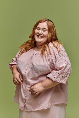 Pembe bluzlu büyük beden bir kadın yeşil arka planda neşeyle güler..