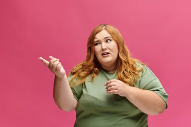 A woman with bright red hair gestures to the side against a vibrant pink background. clipart