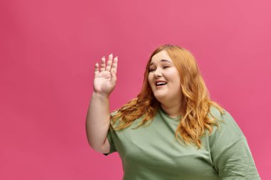 Uzun kızıl saçlı bir kadın gülümsüyor ve el sallıyor. Pembe bir arka plana merhaba diyor..