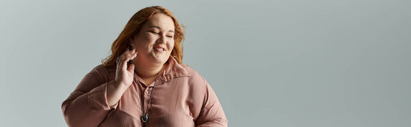 Женщина plus size с рыжими волосами мягко улыбается, в розовой рубашке.