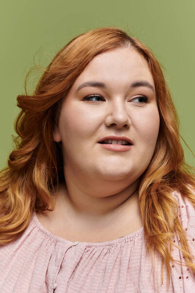 A plus-size woman with red hair looks off-camera, posing in an elegant pink blouse.