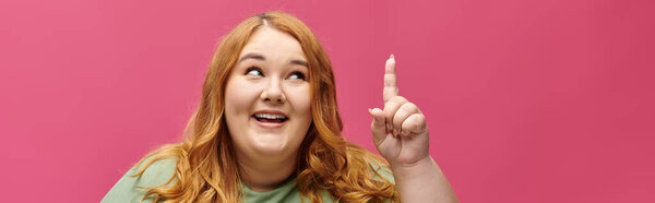 Женщина plus-size с ярко-рыжими волосами улыбается, указывая пальцем вверх.