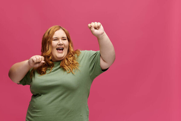 A plus size woman dances enthusiastically in front of a pink backdrop.