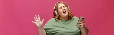 Kızıl saçlı bir kadın müzik eşliğinde şarkı söylüyor. Kulaklık takıyor ve telefon tutuyor..