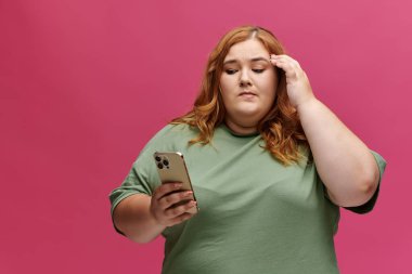 Pembe arka planda telefonu olan yeşil gömlekli kadın düşünceli görünüyor.