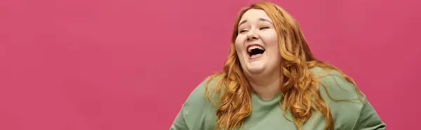 stock image A plus size woman in a green shirt laughs heartily against a pink background.