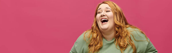 A plus size woman in a green shirt laughs heartily against a pink background.