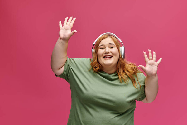 A plus-size woman with red hair smiles and dances, wearing headphones.
