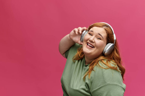 Улыбающаяся женщина с рыжими волосами слушает музыку на розовом фоне в наушниках