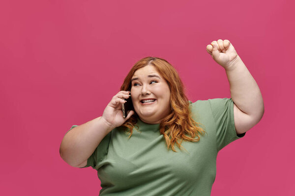 Woman in green shirt smiles, raises fist, talks on phone with pink backdrop