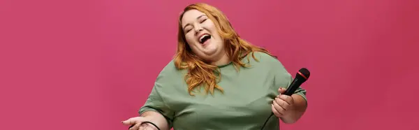 stock image A plus-size woman laughs while holding a microphone, against a pink background.