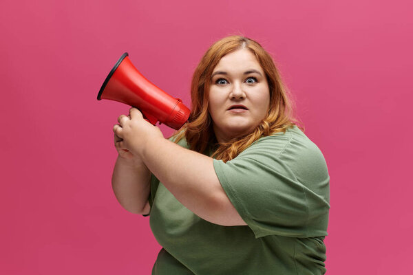 A plus size woman with reddish hair stares intently while shouting through a red megaphone.