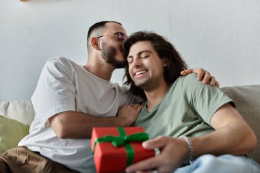 Eşcinsel bir çift koltukta dinleniyor, biri hediye tutarken diğeri kulağına fısıldıyor..