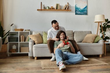 Eşcinsel bir çift koltuğun önünde yere oturur, bir adam diğerine hediye sunar..