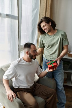 Bir adam partnerine bir hediye verir, o da sandalyeye oturur ve mutlu bir şekilde gülümser..