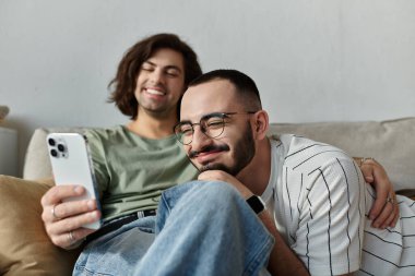 İki adam bir kanepede sarılıyor, biri telefonuna gülüyor, diğeri de ona gülümsüyor.