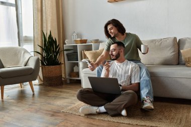 A couple sits on the floor, one man using a laptop while the other looks at his phone. clipart