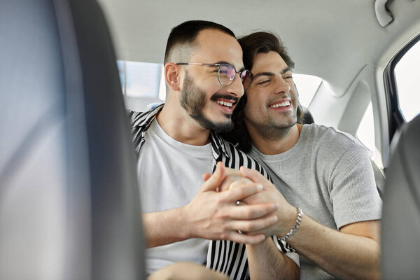 Two men hold hands and smile while sitting in the backseat of a car.