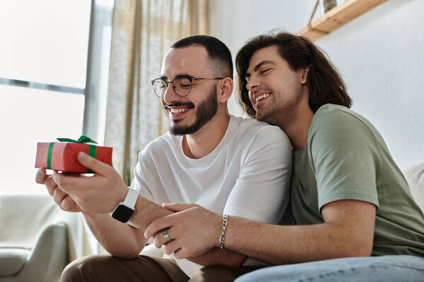A gay couple embraces as one opens a gift, smiling brightly.