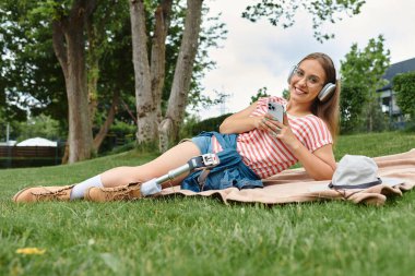 Protez bacaklı genç bir kadın parkta bir battaniyeye uzanıyor, müzik dinliyor ve telefonunu kullanıyor..