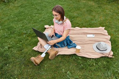Protez bacaklı genç bir kadın parkta dizüstü bilgisayarıyla çalışıyor..