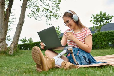 Protez bacaklı genç bir kadın parkta bir battaniyeye oturmuş laptopuyla çalışıyor ve kahvesini yudumluyor..