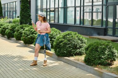 Protez bacaklı genç bir kadın şehir parkında yürüyor, telefonunu kullanıyor ve yaz gününün tadını çıkarıyor..