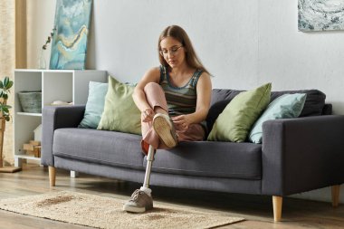 A young woman with a prosthetic leg sits on a couch in her living room, tying her shoe. clipart