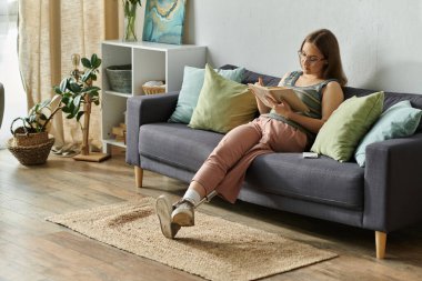 A young woman with a prosthetic leg sits on a couch in her living room, engrossed in a book. clipart