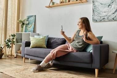 Protez bacaklı genç bir kadın modern oturma odasında bir koltukta oturuyor, gülümsüyor ve telefonunu kullanırken el sallıyor..