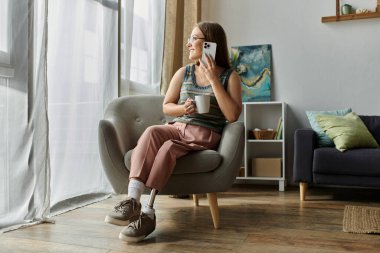 A woman in a living room armchair with prosthetic leg smiles during phone call, holding coffee cup clipart