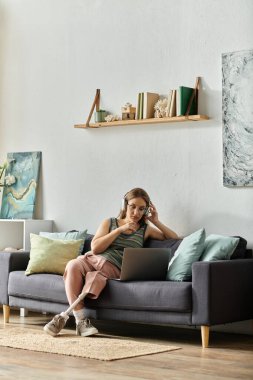 A young woman with a prosthetic leg sits on a couch in her living room, wearing headphones and using a laptop. clipart