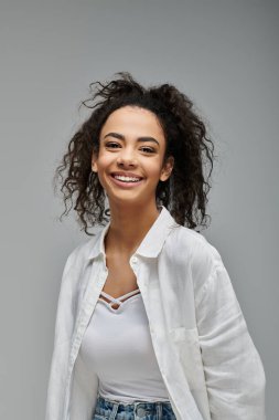 Kıvırcık saçlı genç bir Afrikalı Amerikalı kadın gri bir arka plana doğru gülümsüyor..