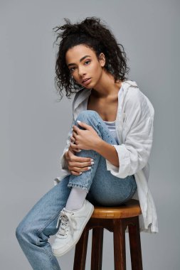 Kıvırcık saçlı genç bir kadın tahta bir taburede oturuyor, beyaz gömlek ve kot pantolon giyiyor..
