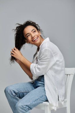 A young African American woman with curly hair sits on a white chair, smiling and playfully touching her hair. clipart