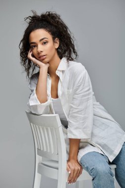 A young woman with curly hair sits on a white chair, resting her chin on her hand, looking thoughtfully at the camera. clipart