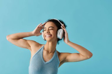 Mavi elbiseli genç bir kadın, kulaklıkla müzik dinlerken mutluluktan gözleri kapalı, parlak mavi bir arka fonda duruyor..
